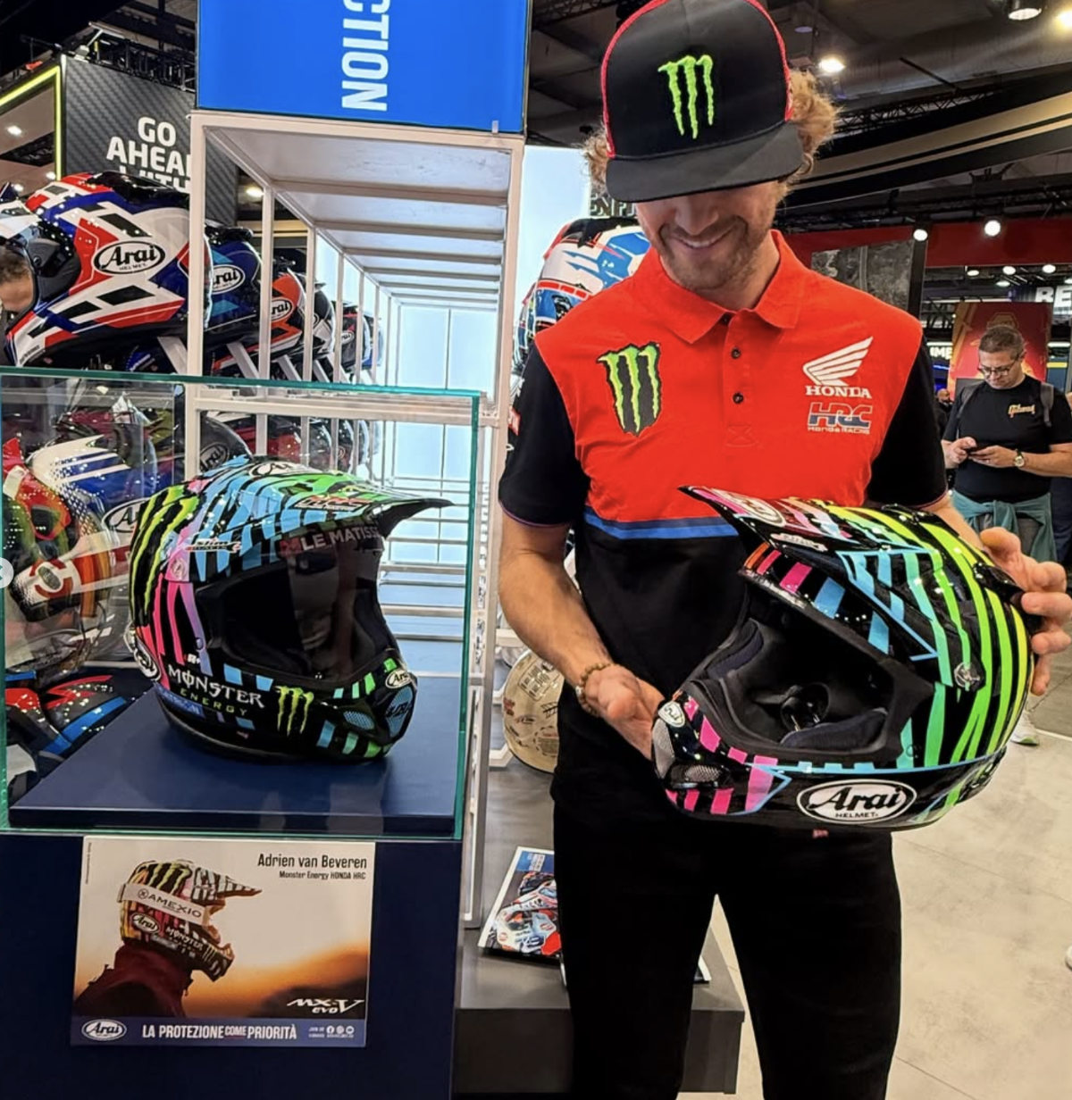 Adrien Van Beveren tenant le casque Replica Officiel Arai X VBA442 tandis que le casque original est exposé en vitrine derrière lui.